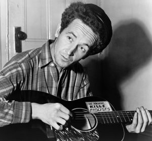 Woody Guthrie with guitar that has sticker on it: "This machine kills fascists"
